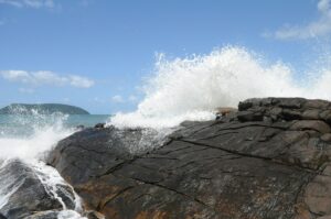 A large, dark gray slab of rock in the foreground, with the ocean and blue sky in the background. A wave crashes on the slab of rock, spraying white foam in the air.