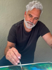 A white man with grey hair and a close-cropped grey beard, wearing a black T-shirt, holds a paintbrush above a canvas painted with abstract blue-green patterns.