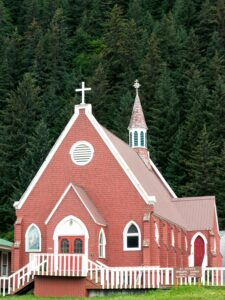 A small church, with red shingles covering its walls and a small white cross atop its a-frame roof, against a backdrop of evergreen trees.