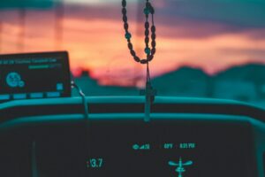 A cross with prayer beads hangs above the center console of a car. All we can see is the clock, a GPS unit, and the cross hanging in the foreground. In the background, through the windshield, we can see the sunrise.